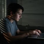 Iemand werkt s nachts op een laptop in een schemerige kamer met jaloezieen, sfeer van privacy en digitaal toezicht