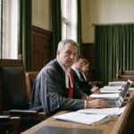 Een rechter in toga kijkt naar de camera achter een bureau in een rustige rechtszaal, met een laptop en documenten voor zich.