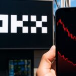 Het OKX-logo op een groot scherm met op de voorgrond een telefoon met een dalende koersgrafiek
